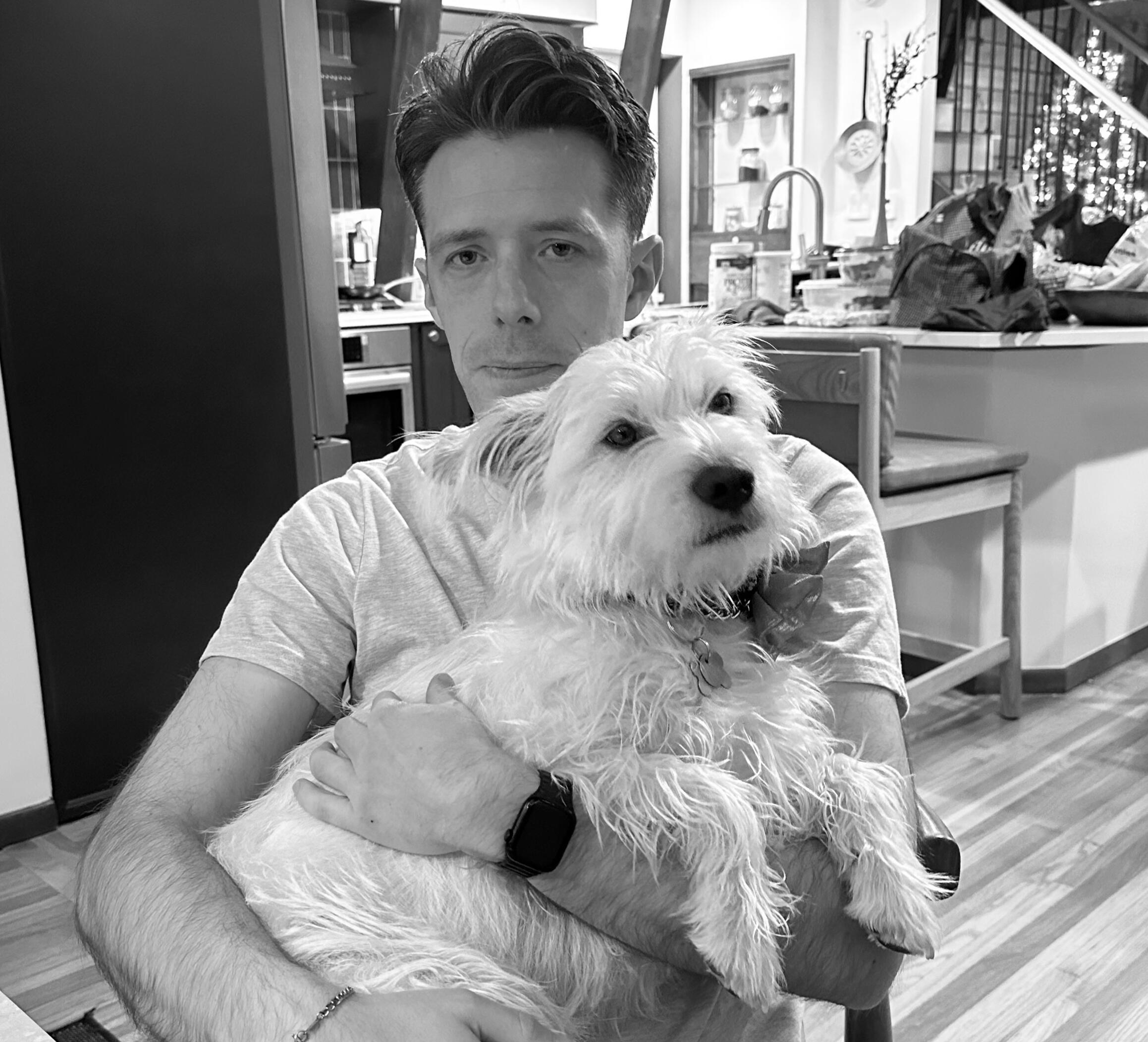 Christian & Monty Caucasian male with tussled hair holding a white scruffy dog in his lap.